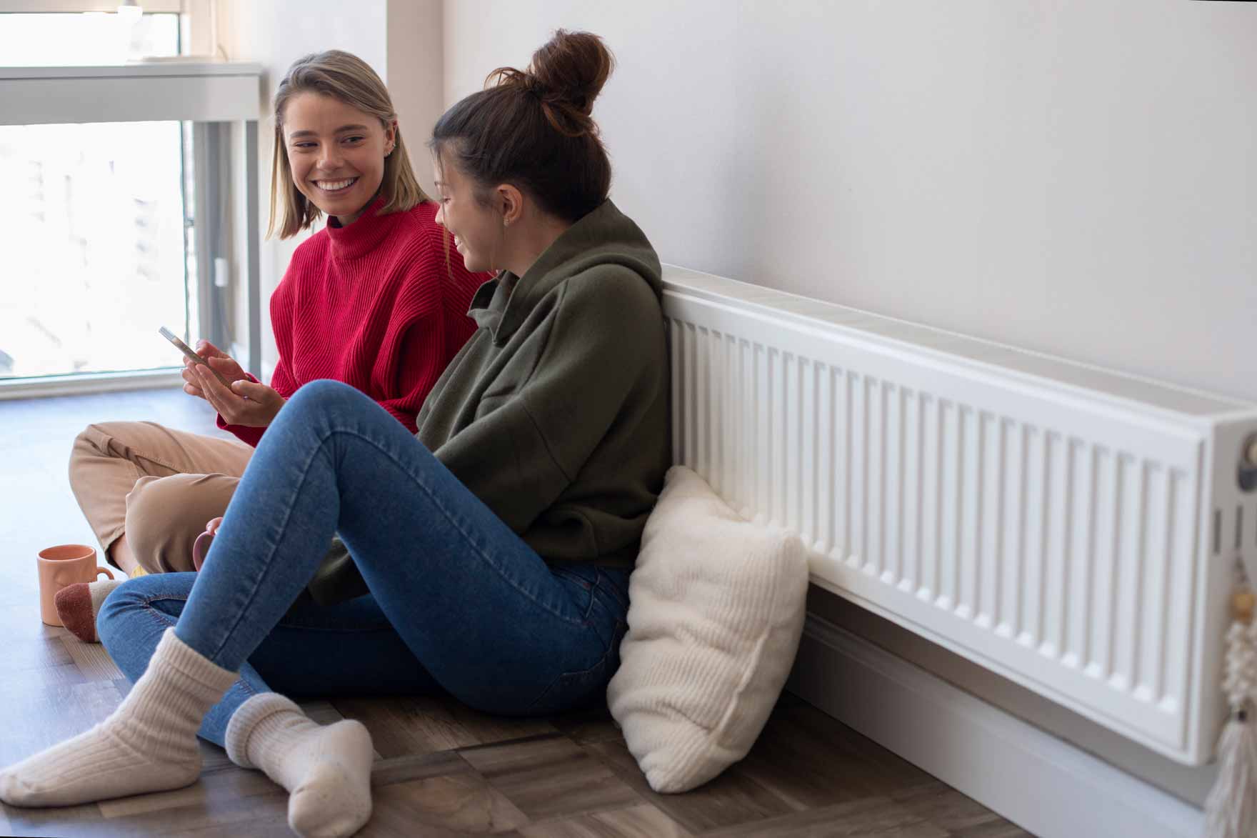 Dos mujeres jóvenes hablando dentro de casa sentadas delante de radiador